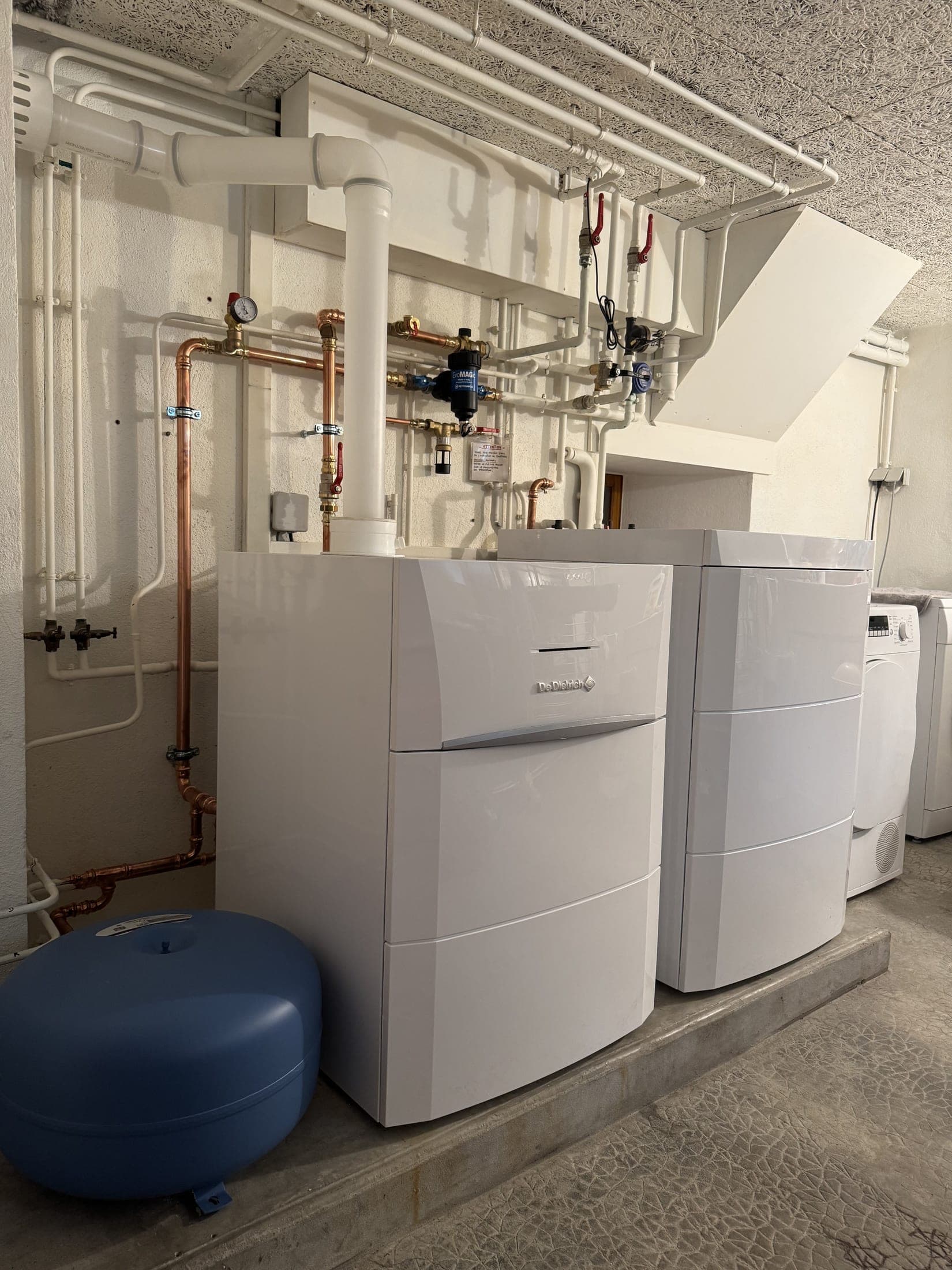 Chaufferie : deux modules blancs sur un socle béton, tuyauteries en cuivre et conduites calorifugées, vase d'expansion bleu.