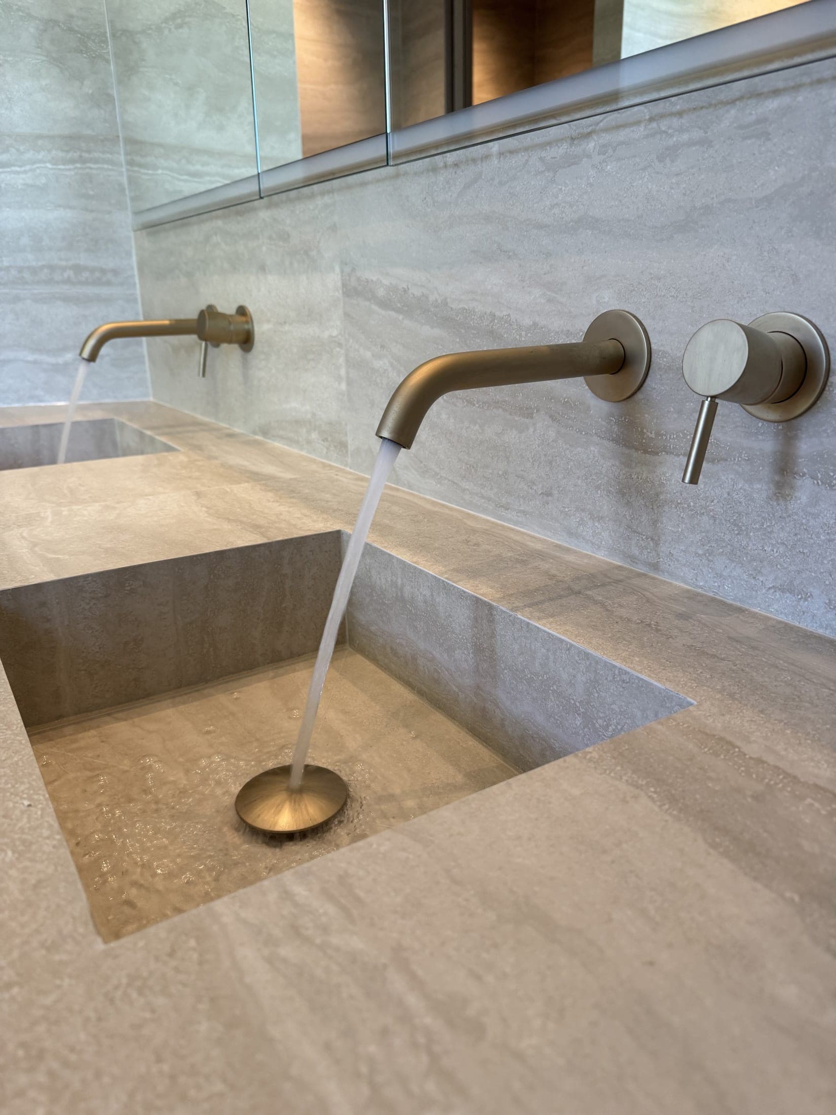 Plan de toilette clair veiné effet pierre, avec deux vasques carrées creusées dans la masse et becs muraux bronze brossé.