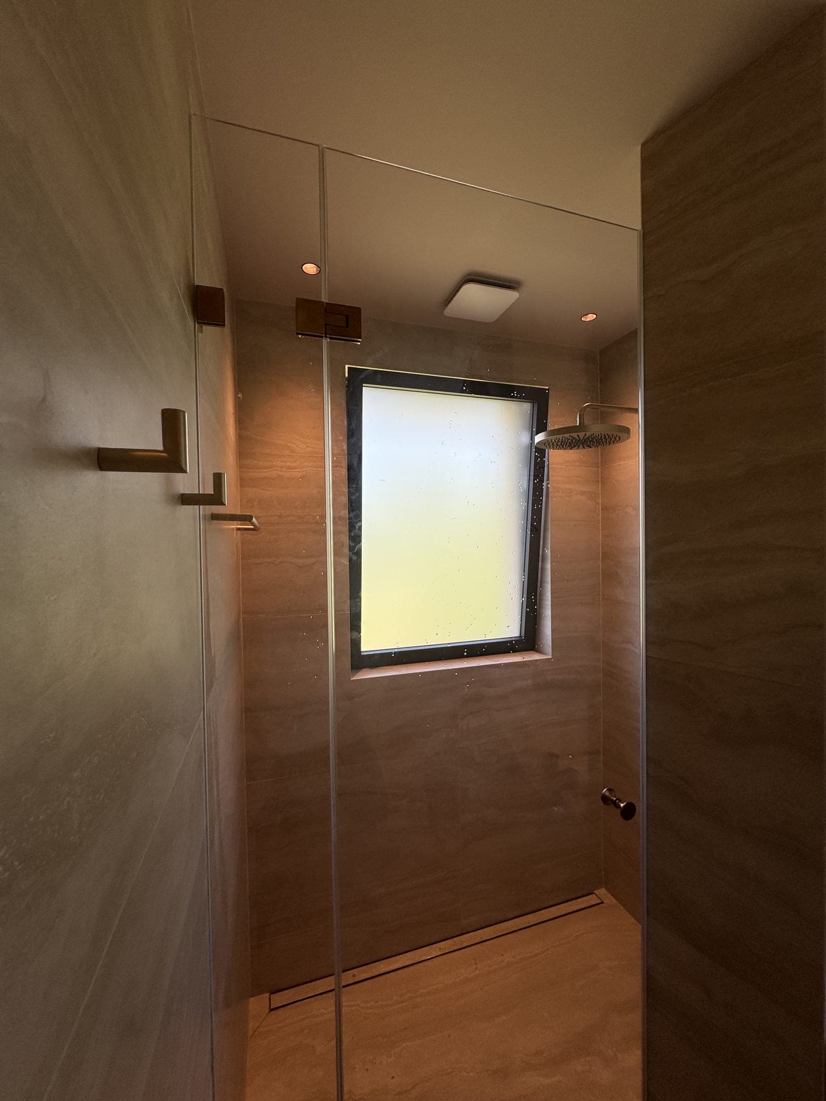 Douche à l'italienne terminée en carrelage effet pierre veinée : pomme de douche dorée, paroi de verre et fenêtre translucide au fond.