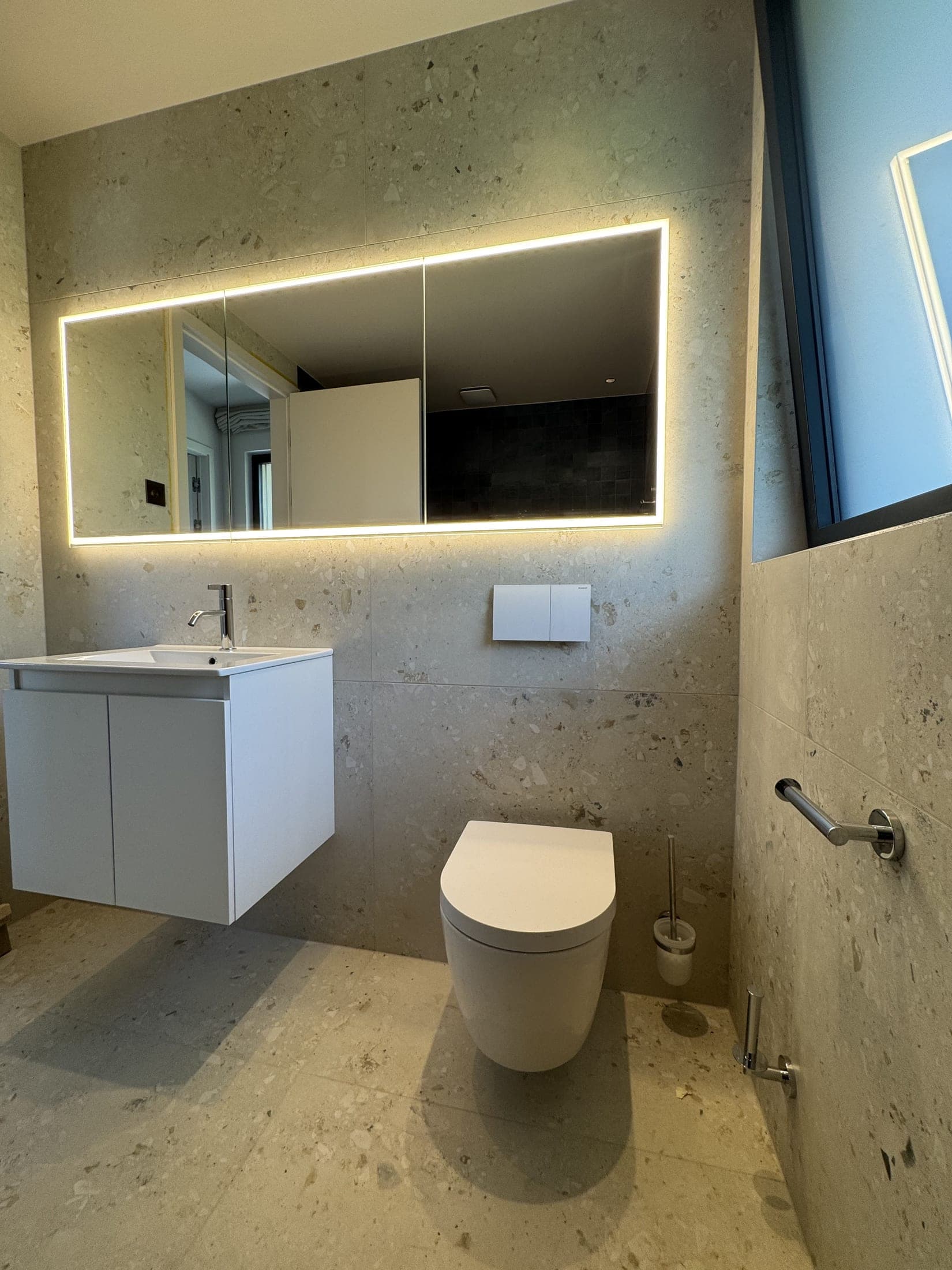 Salle de bain en carrelage gris effet pierre : meuble vasque suspendu, miroir rétroéclairé et WC adossé au mur.