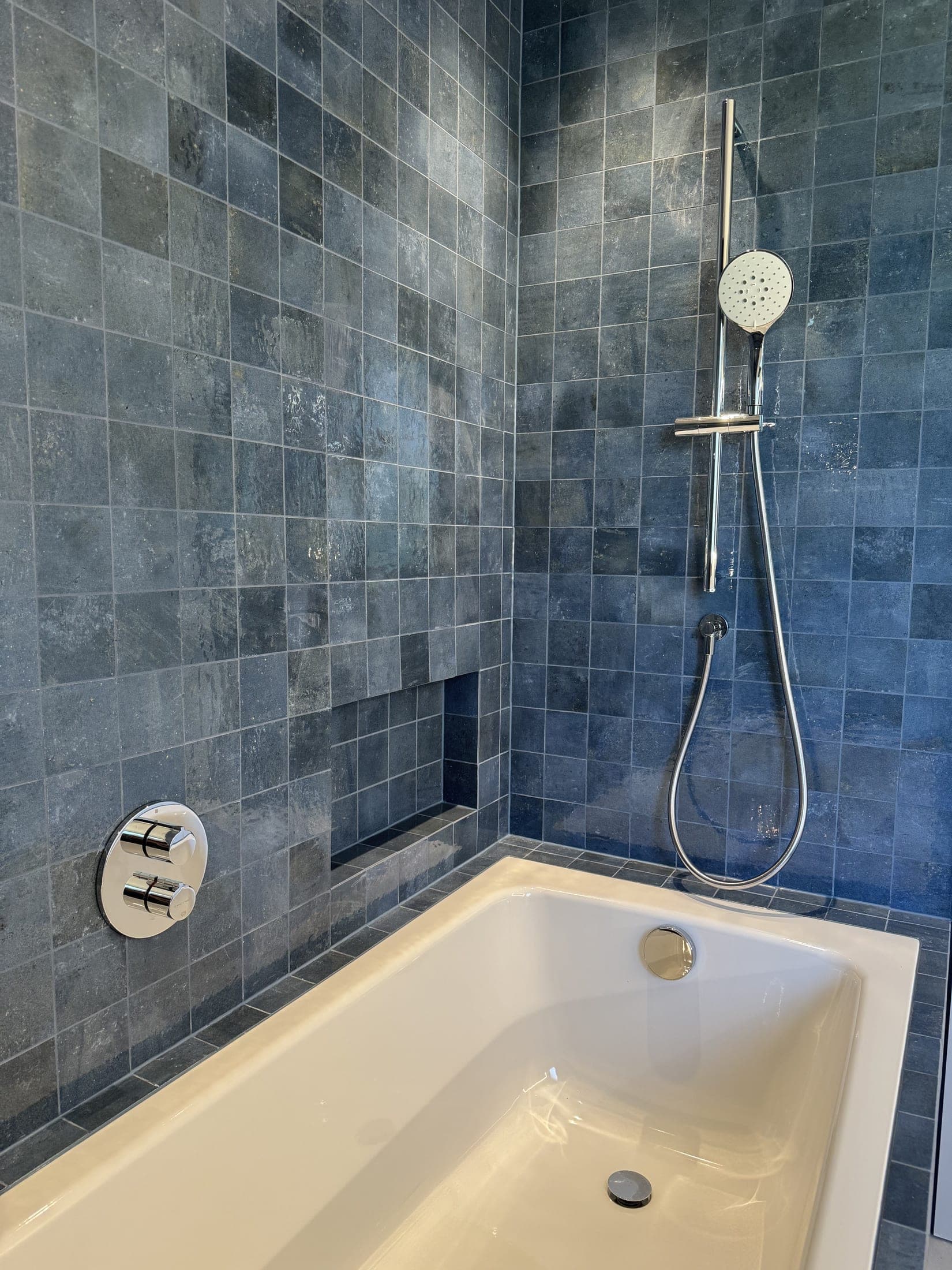 Baignoire blanche entourée de petits carreaux bleus, barre de douche chromée et mitigeur encastré rond à deux commandes.