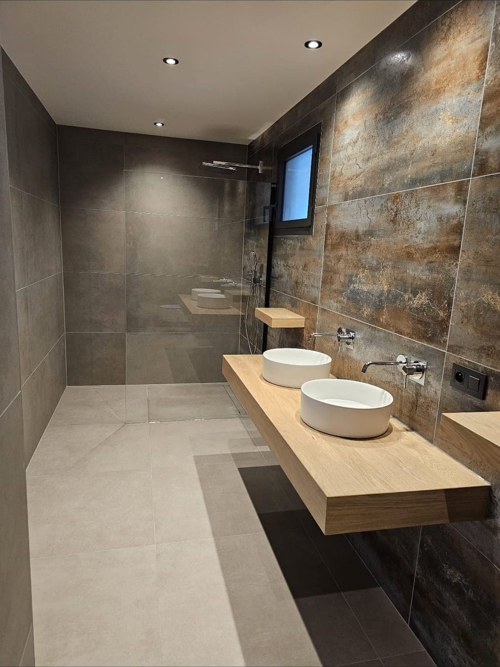 Salle de bain tout en longueur : douche à l'italienne au fond, deux vasques rondes sur tablette en bois et mur effet métal oxydé.