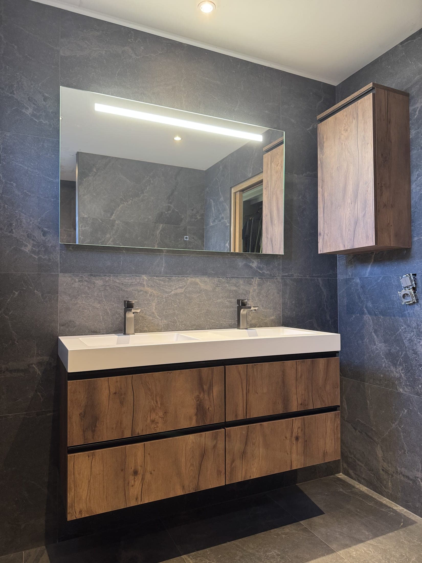 Meuble suspendu à façades effet bois avec plan double vasque, deux mitigeurs chromés et grand miroir éclairé.