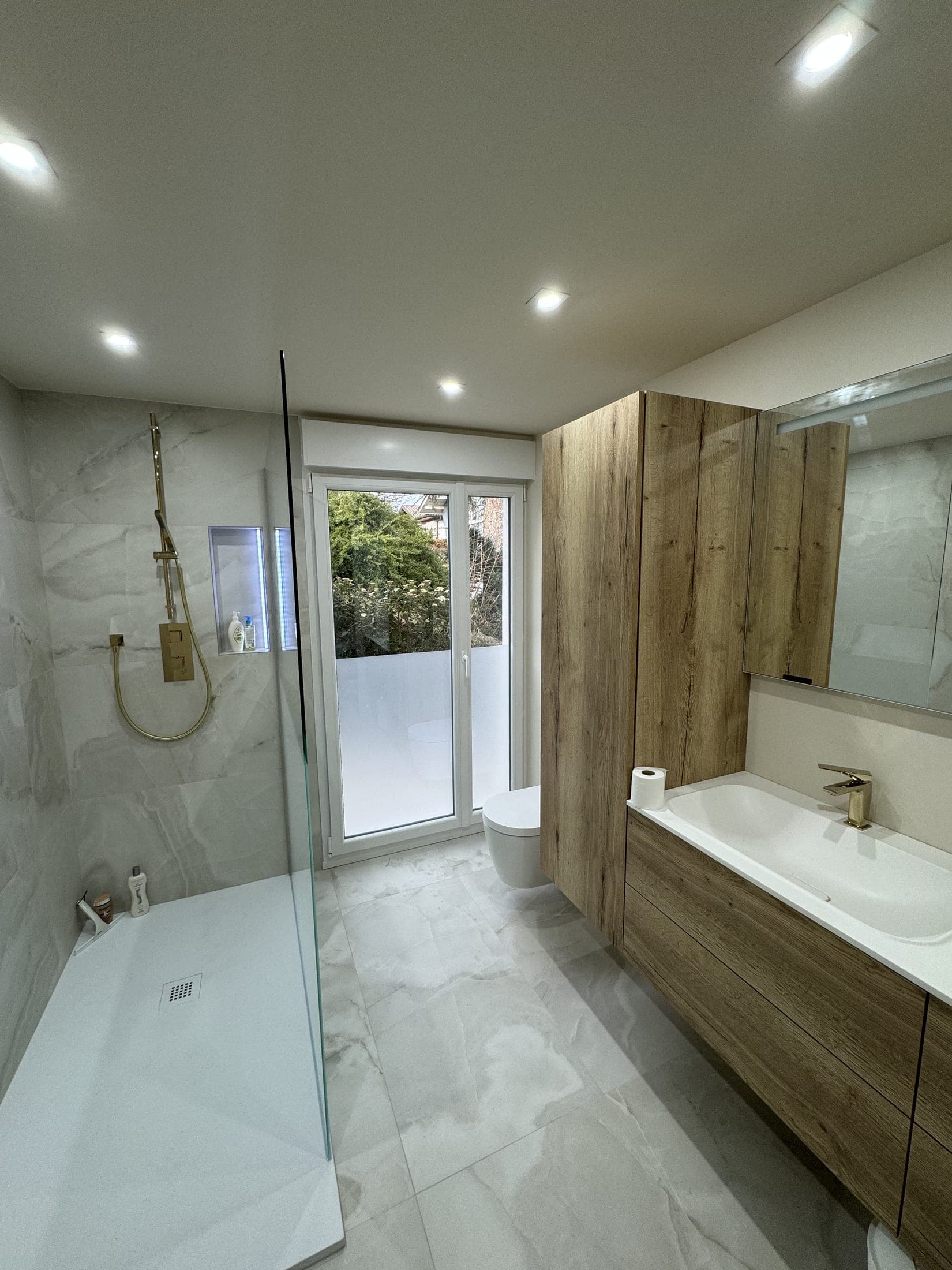 Salle de bain claire : douche à l'italienne avec paroi de verre, WC suspendu et meuble vasque clair, carrelage effet marbre.