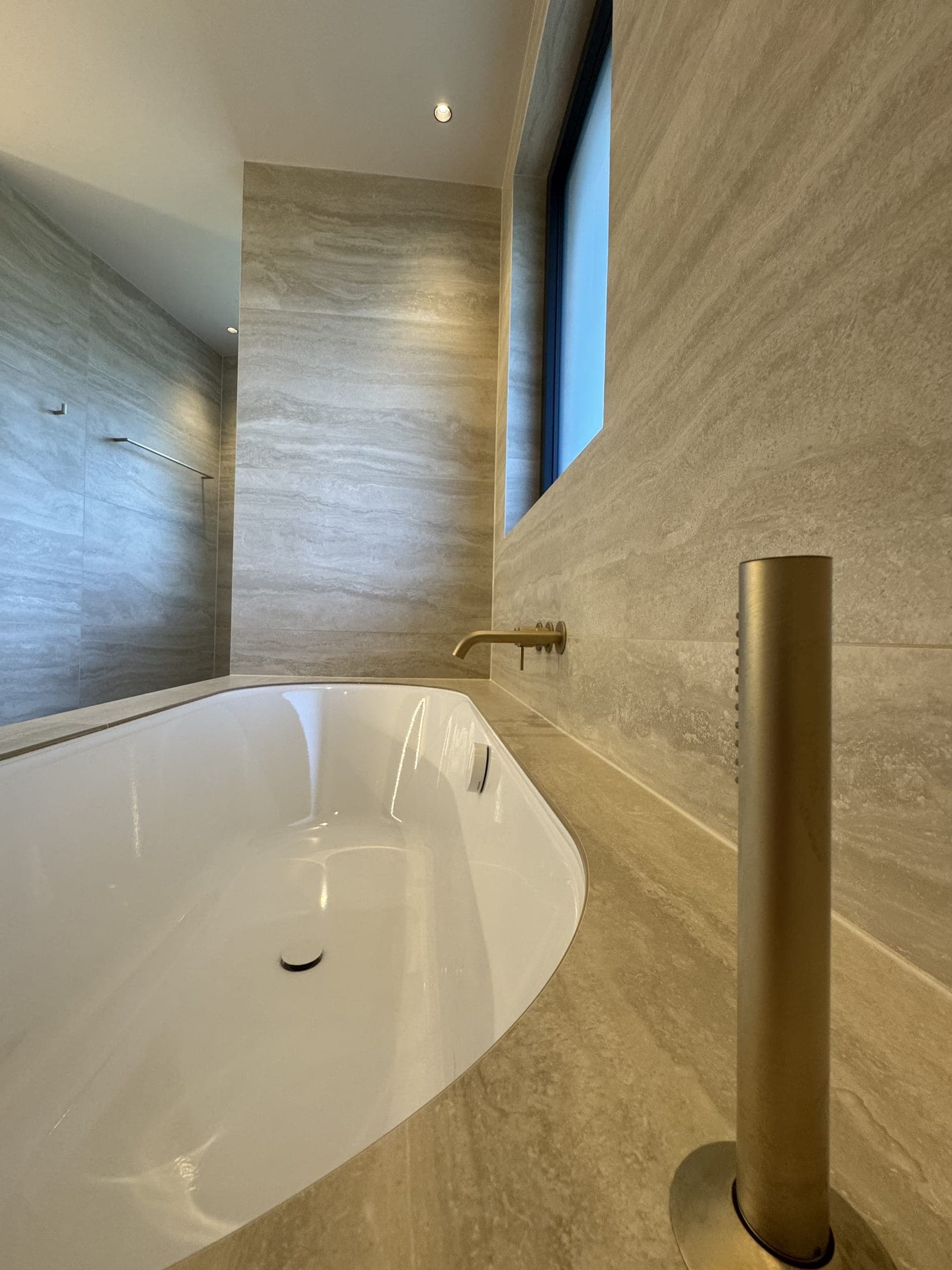 Salle de bain beige : baignoire ovale blanche, bec de remplissage mural doré et colonne de douchette dorée.