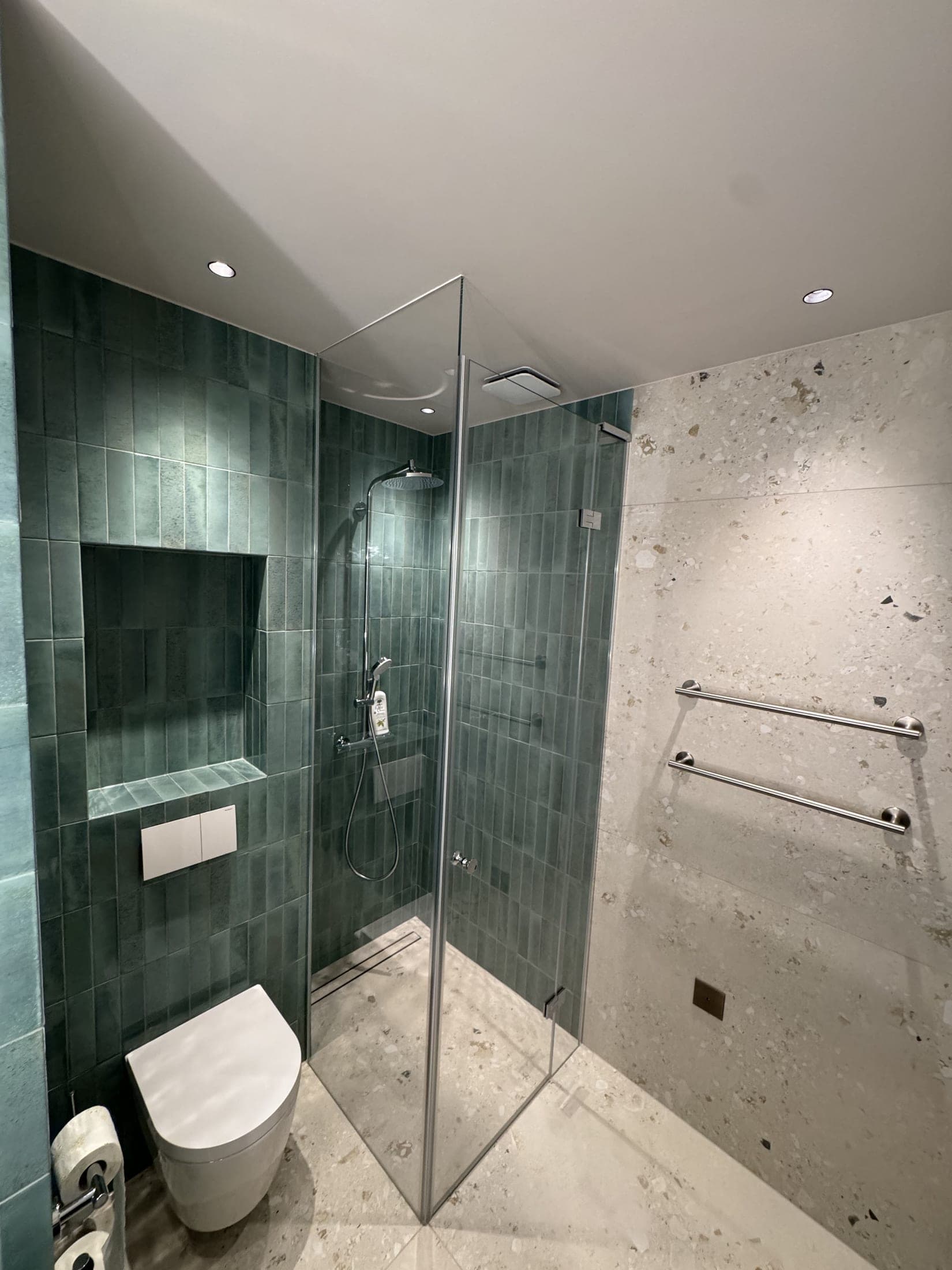 Salle de bain en carrelage vert : douche d'angle vitrée porte ouverte, niche carrelée, WC suspendu et mur en pierre claire.