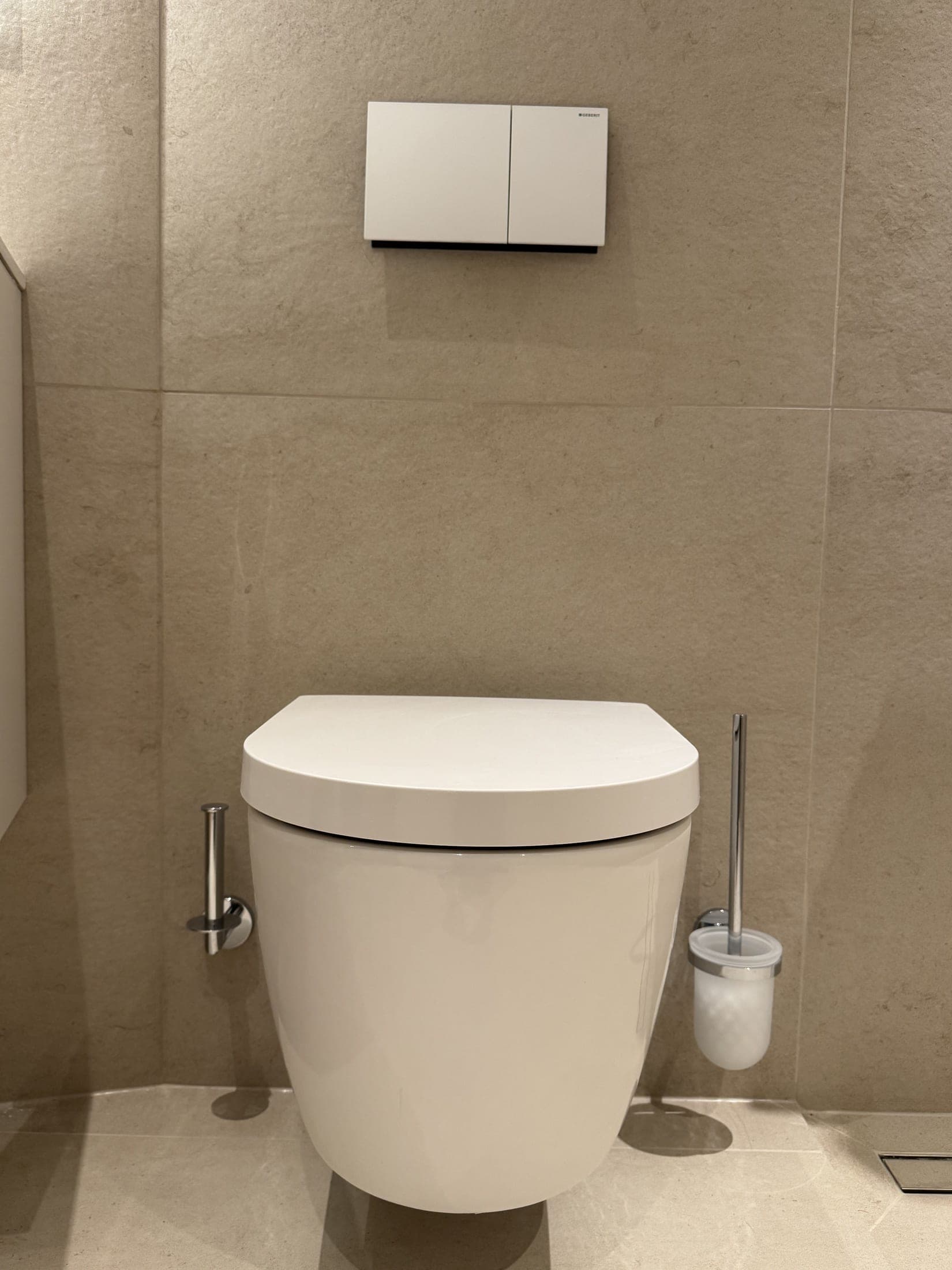 WC suspendu blanc sur mur en carrelage beige grand format, plaque de commande à deux touches et porte-rouleau chromé.